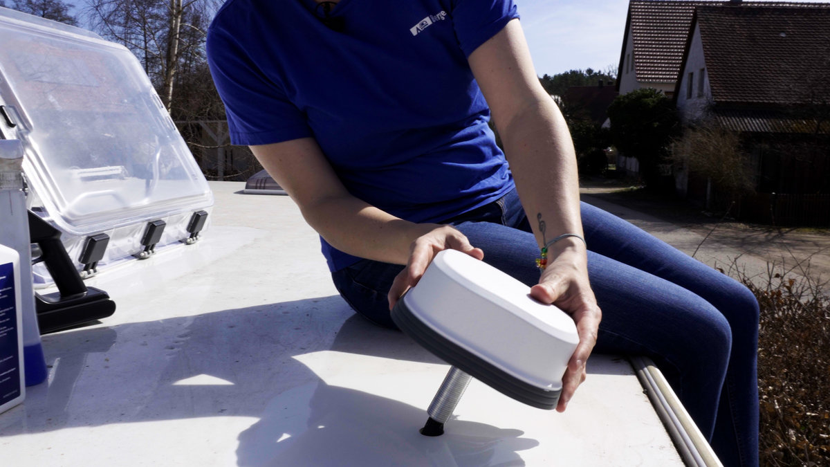 Person sitzt auf einer wei&szlig;en flachen Oberfl&auml;che, nur Oberk&ouml;rper und Arme sichtbar, h&auml;lt ein wei&szlig;es abgerundetes rechteckiges Ger&auml;t mit kurzem Metallstift; offene Kunststoffbox und H&auml;user im Hintergrund