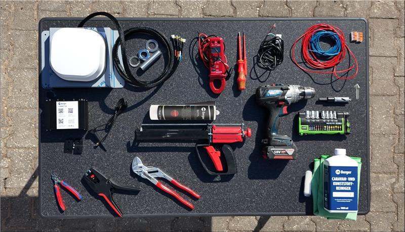 Verschiedene Werkzeuge und Kabel auf einer schwarzen Matte: wei&szlig;e runde Box, aufgewickeltes schwarzes Kabel, drei Metallringe, rote Kabel mit Klemmen, aufgewickeltes blaues Kabel, schwarzes rechteckiges Ger&auml;t, zwei Kartuschen mit Kartuschenpressen, A