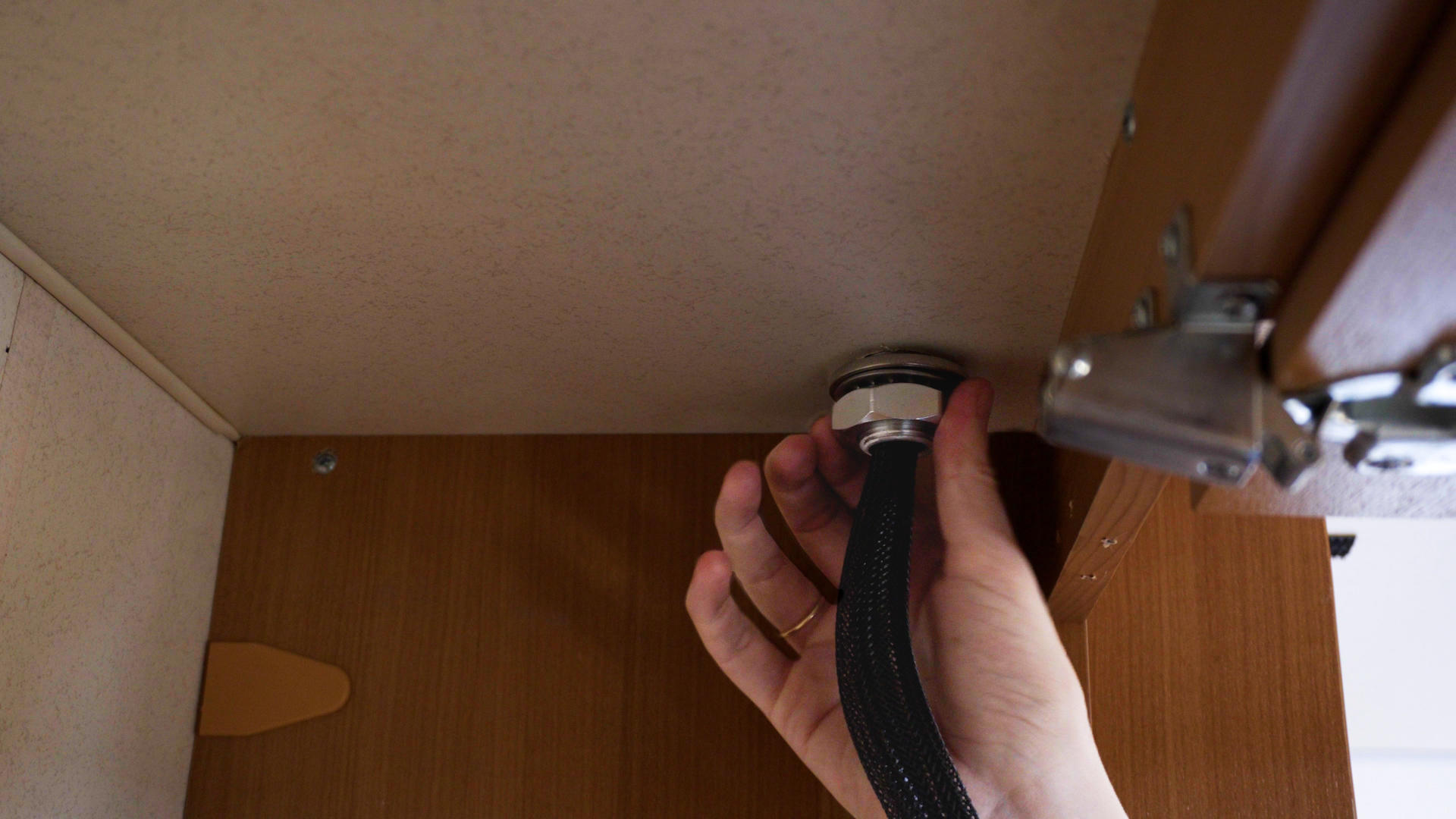 Hand h&auml;lt ein B&uuml;ndel schwarzer Kabel, das durch eine runde Kabeldurchf&uuml;hrung in der Oberseite eines Holzschranks f&uuml;hrt; rechts ist ein Metall-Scharnier sichtbar