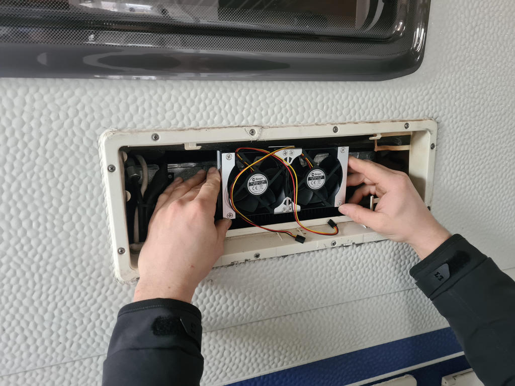 Zwei kleine L&uuml;fter in einem rechteckigen Einbauschacht, zwei H&auml;nde halten die L&uuml;fter, sichtbare Kabel und Schrauben, wei&szlig;e strukturierte Wand