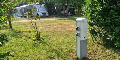 Wei&szlig;er Versorgungspoller mit Steckdosen im Vordergrund; dahinter ein Wohnmobil mit ausgefahrener Markise auf einer Rasenfl&auml;che; B&auml;ume und Str&auml;ucher