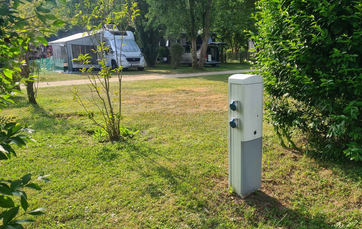 Wei&szlig;er Versorgungspoller mit Steckdosen im Vordergrund; dahinter ein Wohnmobil mit ausgefahrener Markise auf einer Rasenfl&auml;che; B&auml;ume und Str&auml;ucher