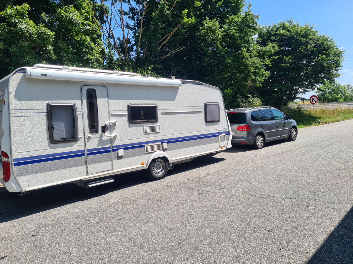 Graues Auto mit wei&szlig;em Wohnwagen am Stra&szlig;enrand unter B&auml;umen, blauer Himmel