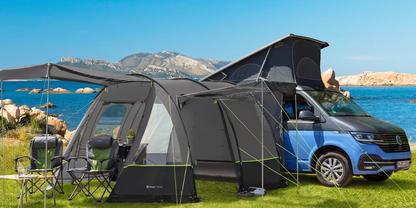 Blauer Camper-Van mit angebautem schwarzem Vorzelt auf gr&uuml;nem Gras am Meeresufer, zwei Campingst&uuml;hle und kleiner Tisch mit Tassen, Felsen und Berge im Hintergrund unter blauem Himmel