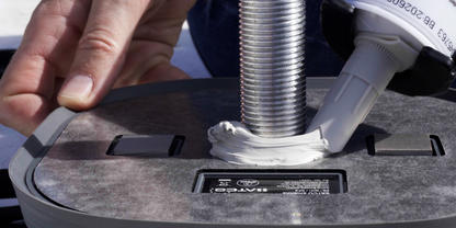 Eine Kartuschenpistole gibt wei&szlig;e Dichtmasse um einen Metallgewindebolzen auf einer grauen Kunststoffbasis ab; eine Hand h&auml;lt die Basis