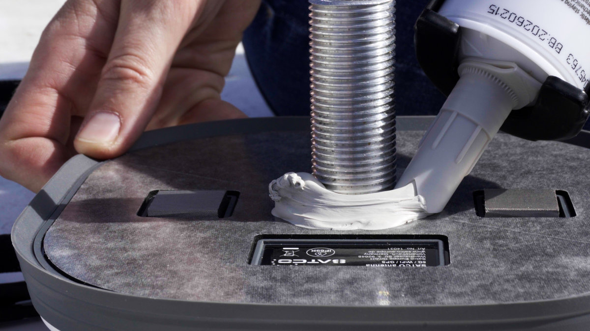 Eine Kartuschenpistole gibt wei&szlig;e Dichtmasse um einen Metallgewindebolzen auf einer grauen Kunststoffbasis ab; eine Hand h&auml;lt die Basis