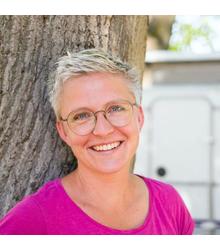 Persona con cabello rubio corto y gafas redondas, viste una camiseta rosa, apoyada sonriente en un &aacute;rbol; fondo claro desenfocado