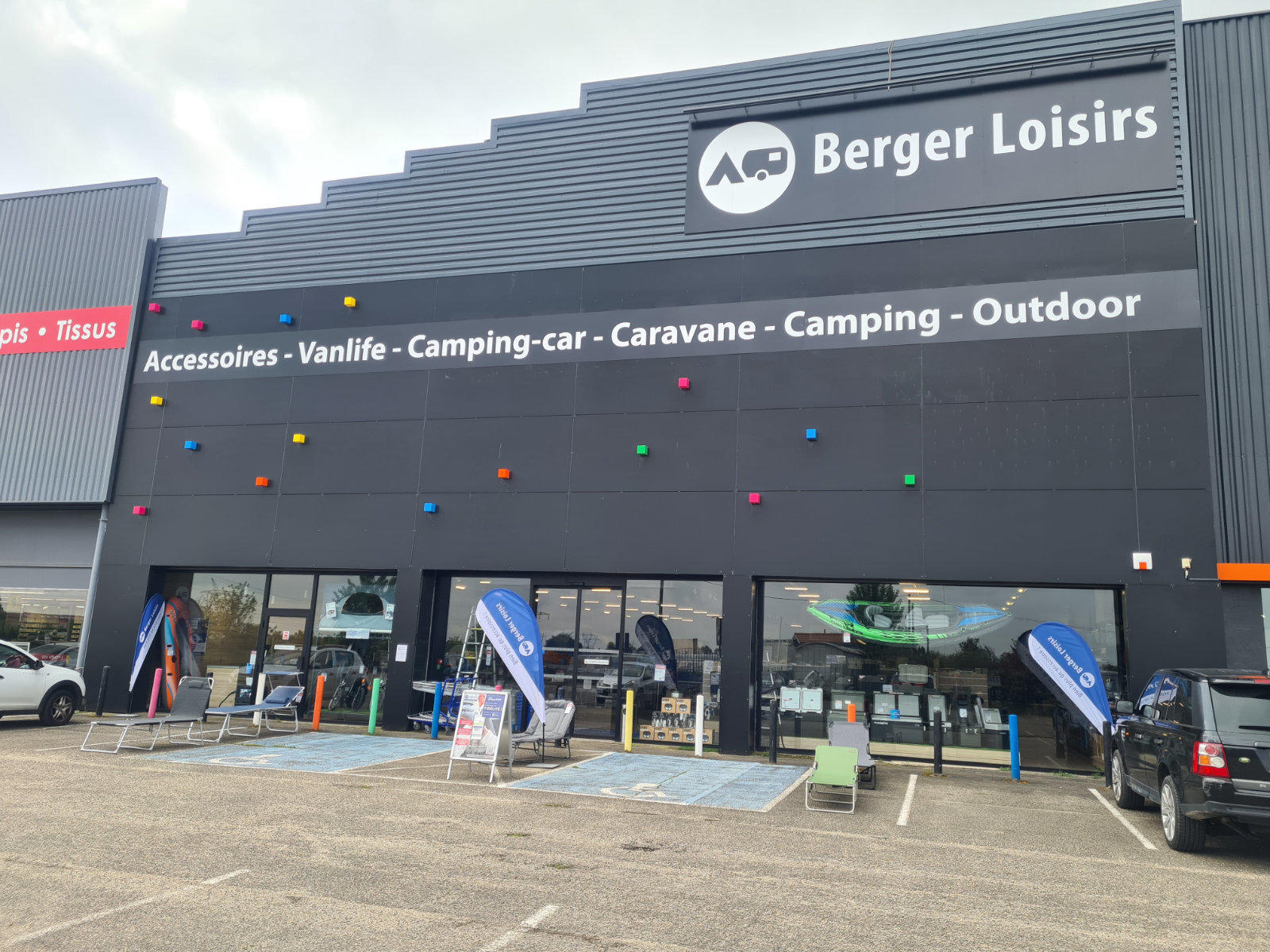 Dunkelgraue Ladenfassade mit gro&szlig;em Schriftzug 'Berger Loisirs', wei&szlig;e Zeile 'Accessoires - Vanlife - Camping-car - Caravane - Camping - Outdoor', bunte runde Punkte an der Fassade, Glaseingang mit blauen Werbefahnen, parkende Autos, bew&ouml;lkter Himmel
