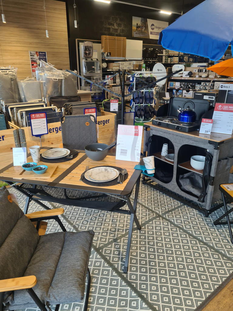 Zwei graue Klappst&uuml;hle mit Holzarmlehnen, ein klappbarer Holztisch mit zwei gedeckten Pl&auml;tzen (Teller, Sch&uuml;ssel, Besteck), ein blauer Sonnenschirm oben rechts, Regale und Kartons mit T&ouml;pfen, Pfannen und K&uuml;chenutensilien im Hintergrund, grauer Teppich