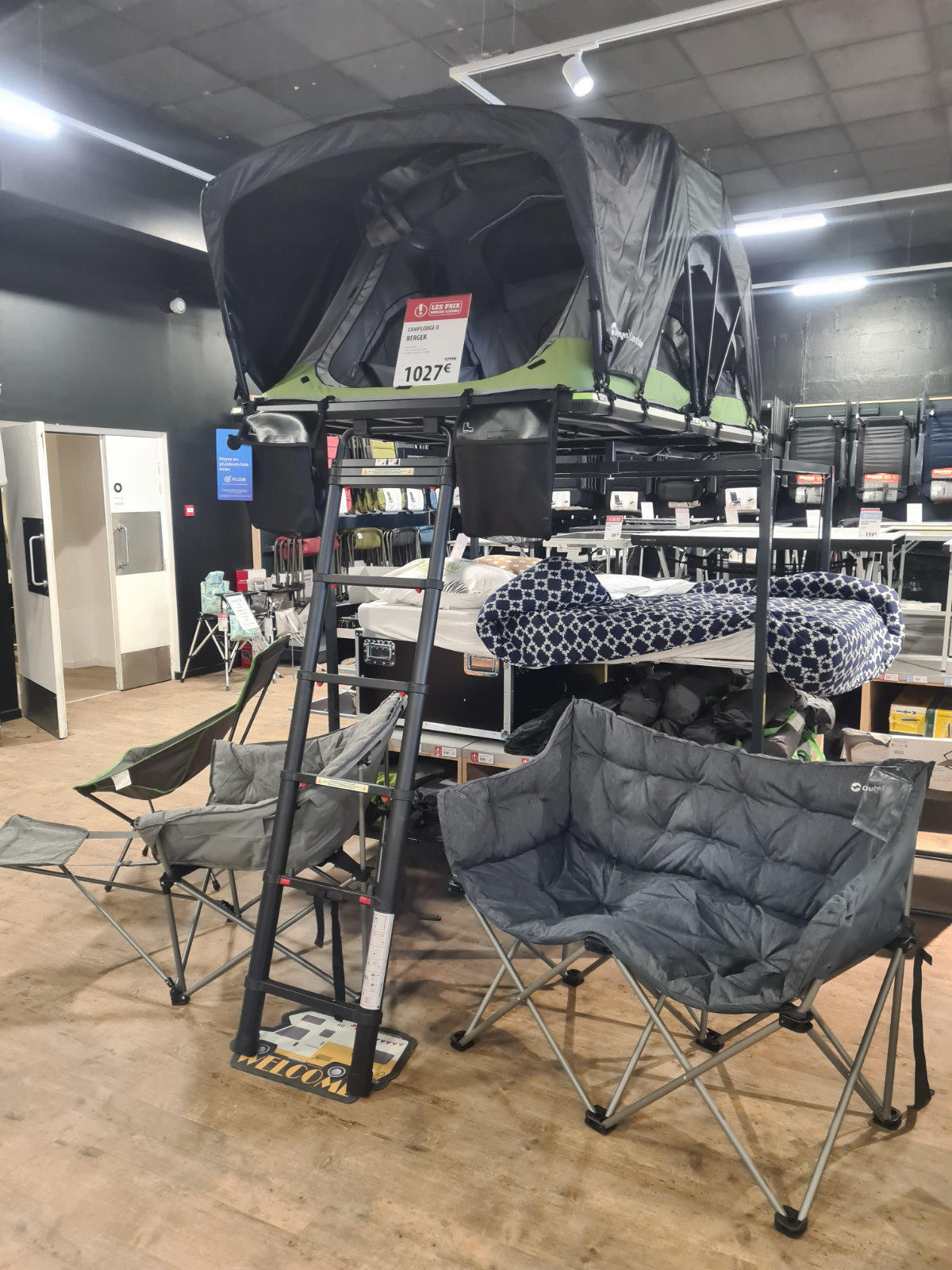 Aufgeklapptes Dachzelt auf Metallgestell mit Leiter, davor zwei graue gepolsterte Klappst&uuml;hle und eine gemusterte Matratze, Preisschild und Regale im Gesch&auml;ft