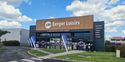 Geb&auml;udefassade mit gro&szlig;em Schriftzug 'Berger Loisirs', Glasfront, zwei stehende Werbefahnen vor dem Eingang, gepflasterte Zufahrt, Rasenfl&auml;che und wolkiger blauer Himmel