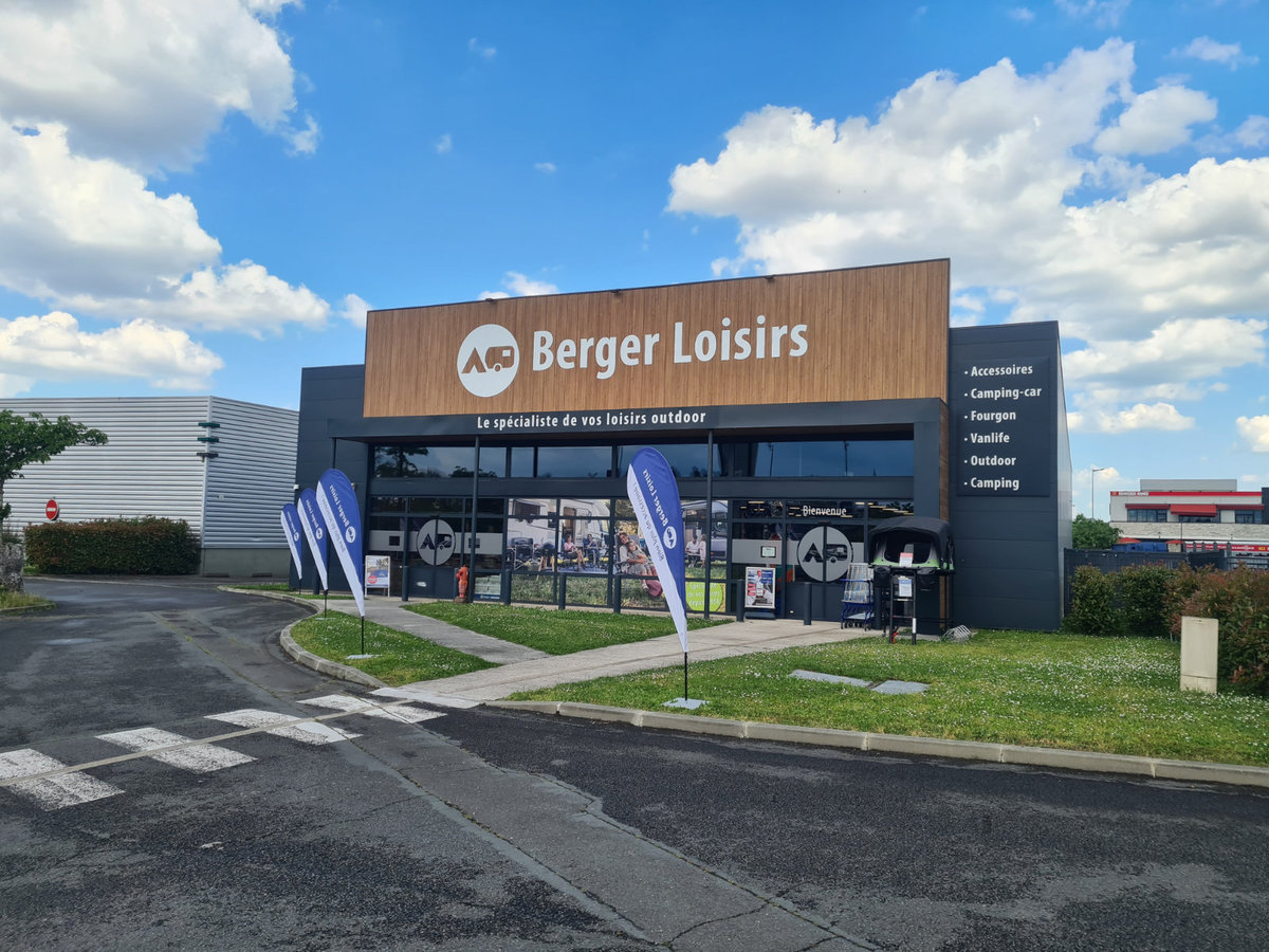 Geb&auml;udefassade mit gro&szlig;em Schriftzug 'Berger Loisirs', Glasfront, zwei stehende Werbefahnen vor dem Eingang, gepflasterte Zufahrt, Rasenfl&auml;che und wolkiger blauer Himmel