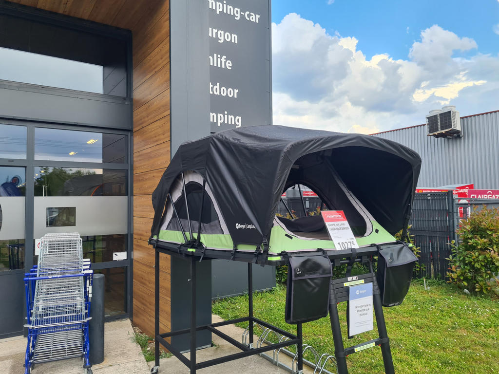 Schwarzes aufgeklapptes Zelt mit gr&uuml;nem Rand auf einem schwarzen Metallgestell vor einem Gesch&auml;ftsgeb&auml;ude; zwei Schilder am Zelt, Einkaufswagen links, Holzverkleidung an der Fassade, Zaun und Nebengeb&auml;ude rechts, blauer Himmel mit Wolken