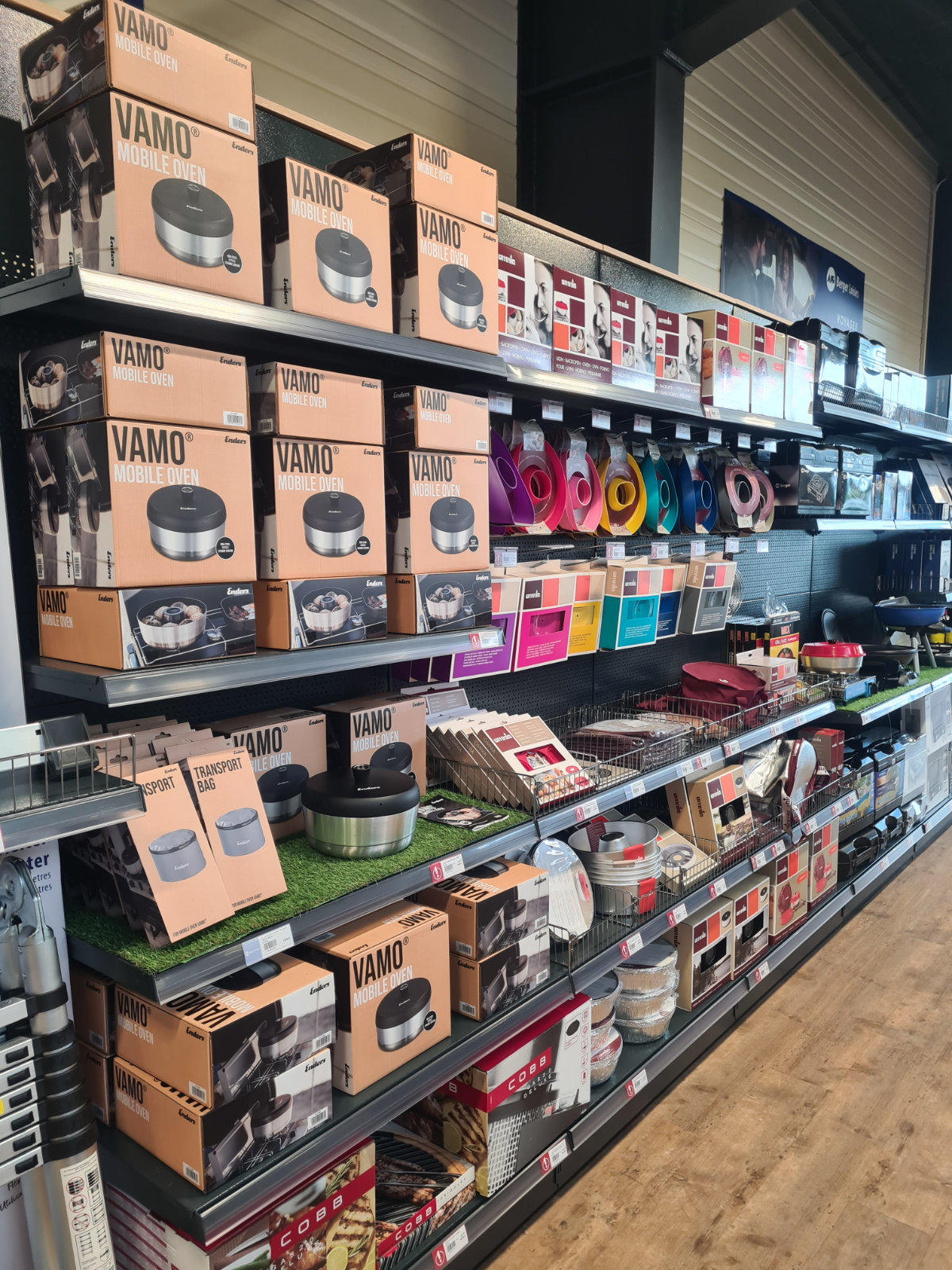 Verkaufsregal mit Kartons und ausgestelltem Kochgeschirr, Pfannen, bunten K&uuml;chenutensilien, Preisschildern und Holzfu&szlig;boden