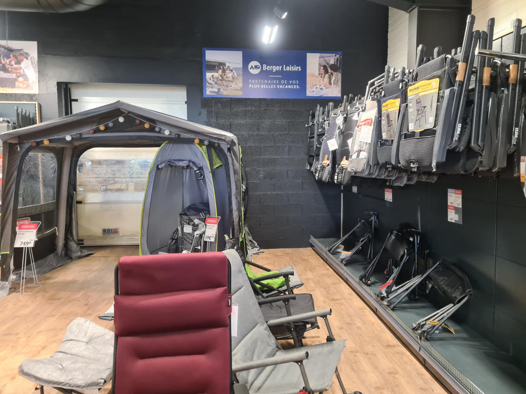 Verkaufsraum mit offenem Campingzelt/Pavillon links, mehreren Campingst&uuml;hlen vorne (eins rot), zusammengeklappten St&uuml;hlen an der rechten Wand, Holzfu&szlig;boden und dunkler R&uuml;ckwand mit Schild