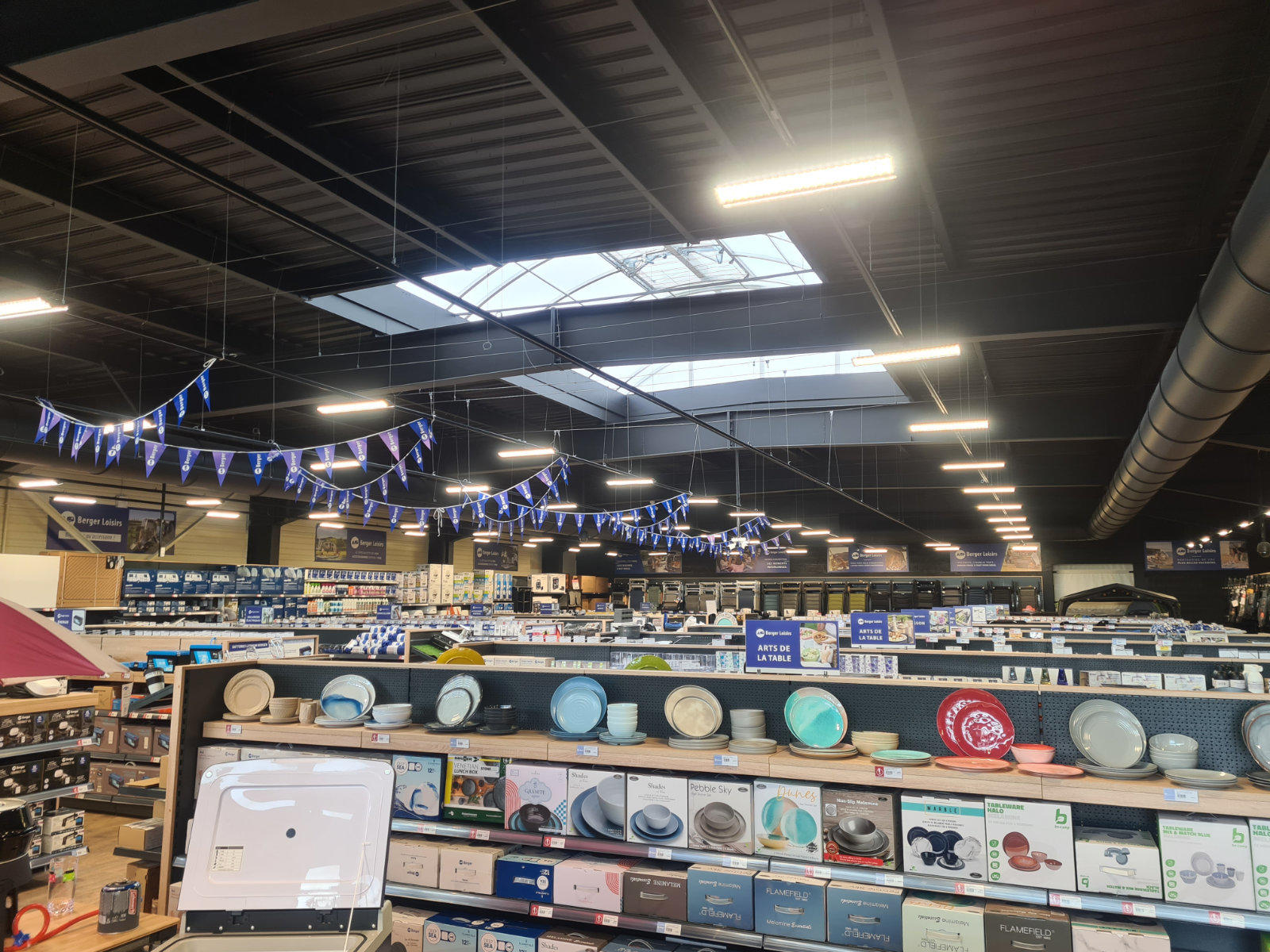 Innenansicht eines gro&szlig;en Gesch&auml;fts mit Regalen voller Teller und Kartons, h&auml;ngenden Leuchten, blauen Wimpelgirlanden, Dachfenstern und sichtbarer Metalldecke mit L&uuml;ftungsrohr