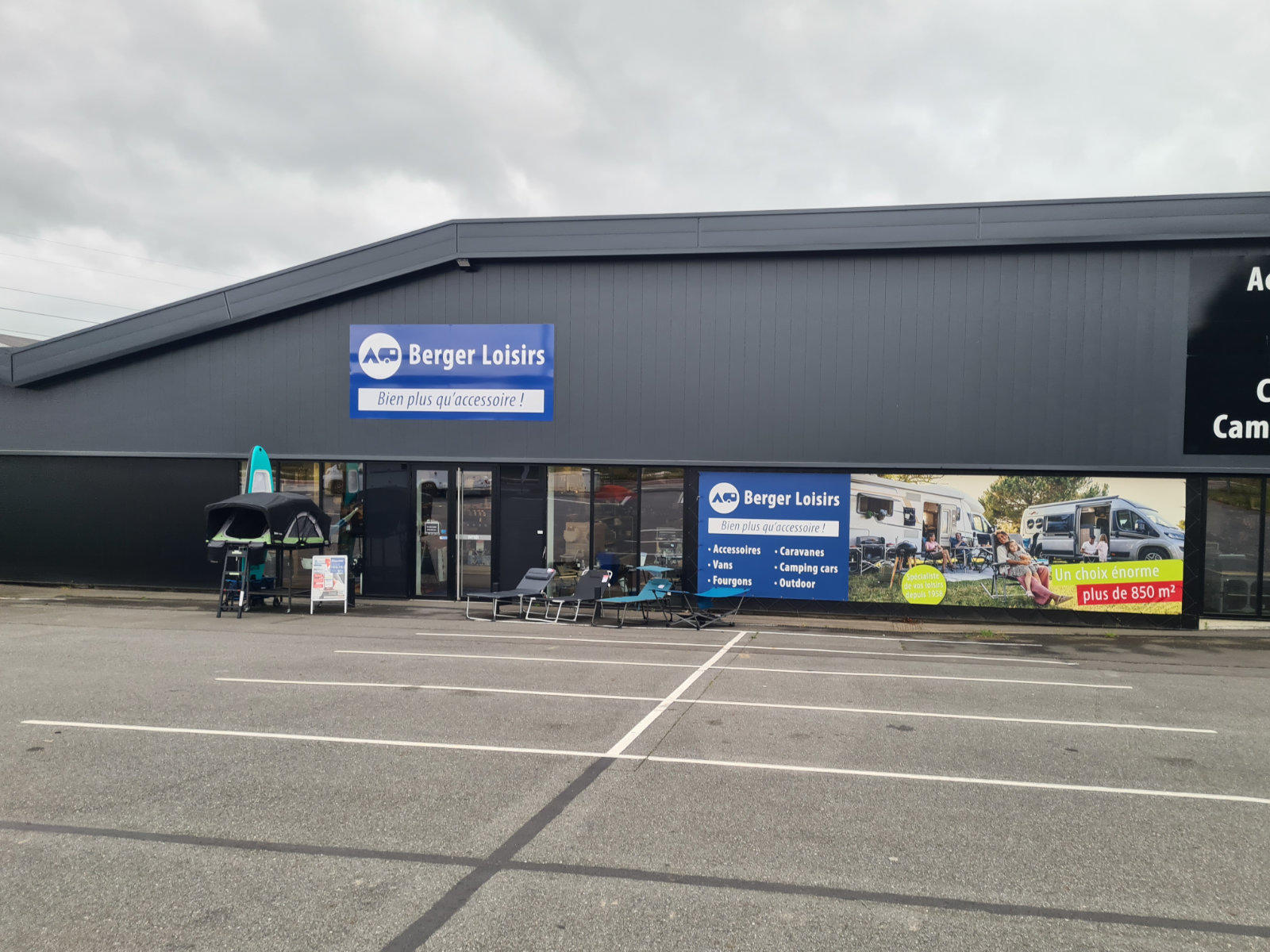 Fassade eines dunkelgrauen Gesch&auml;fts mit blauem "Berger Loisirs"-Schild, Glaseingang, Au&szlig;enaufsteller mit Sonnenschirm und St&uuml;hlen, leerer Parkplatz und bew&ouml;lkter Himmel
