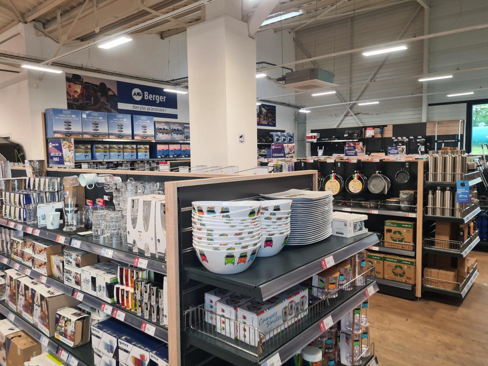 Innensicht eines Haushaltswarenladens mit Regalen; im Vordergrund gestapelte Kunststoffsch&uuml;sseln und wei&szlig;e Teller auf einem Regal, daneben K&uuml;chenutensilien und Besteck; Pfannen an der rechten Wand; Deckenleuchten