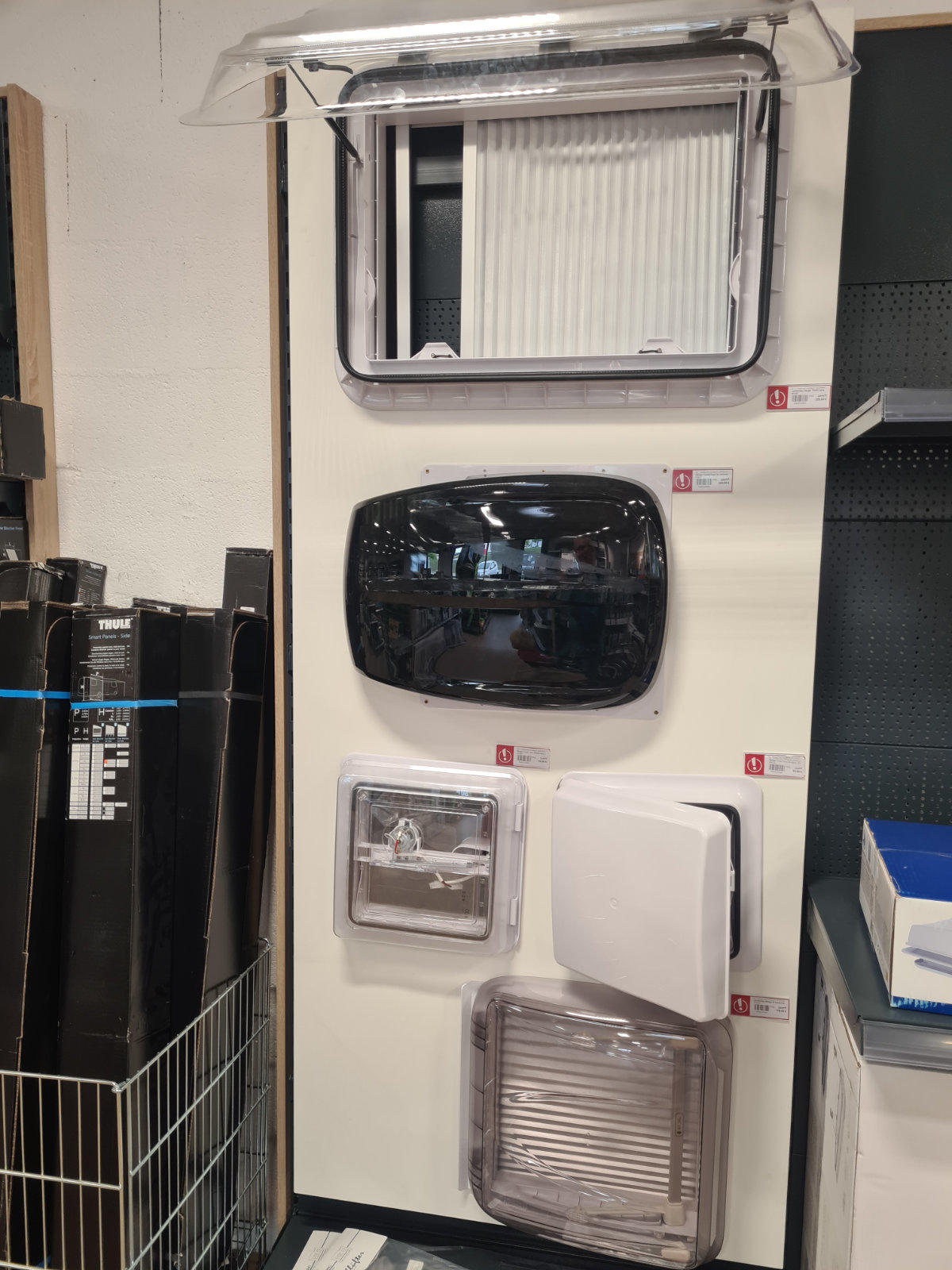 Wei&szlig;e Wand mit f&uuml;nf Kunststoffabdeckungen in rechteckigen und ovalen Formen; links ein Drahtkorb mit aufgerollten schwarzen Rollen; rechts ein Regal mit blauen Kartons