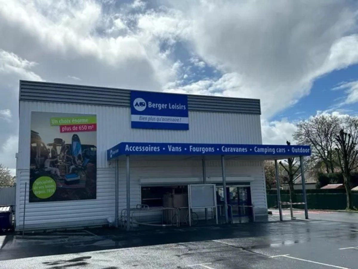 Fassade eines Gesch&auml;ftsgeb&auml;udes mit blauem 'Berger Loisirs'-Schild &uuml;ber dem Eingang, blaues Vordach mit wei&szlig;er Schrift 'Accessoires - Vans - Fourgons - Caravanes - Camping cars - Outdoor', gro&szlig;es Werbeplakat links, Glast&uuml;ren am Eingang, nasser Parkpl