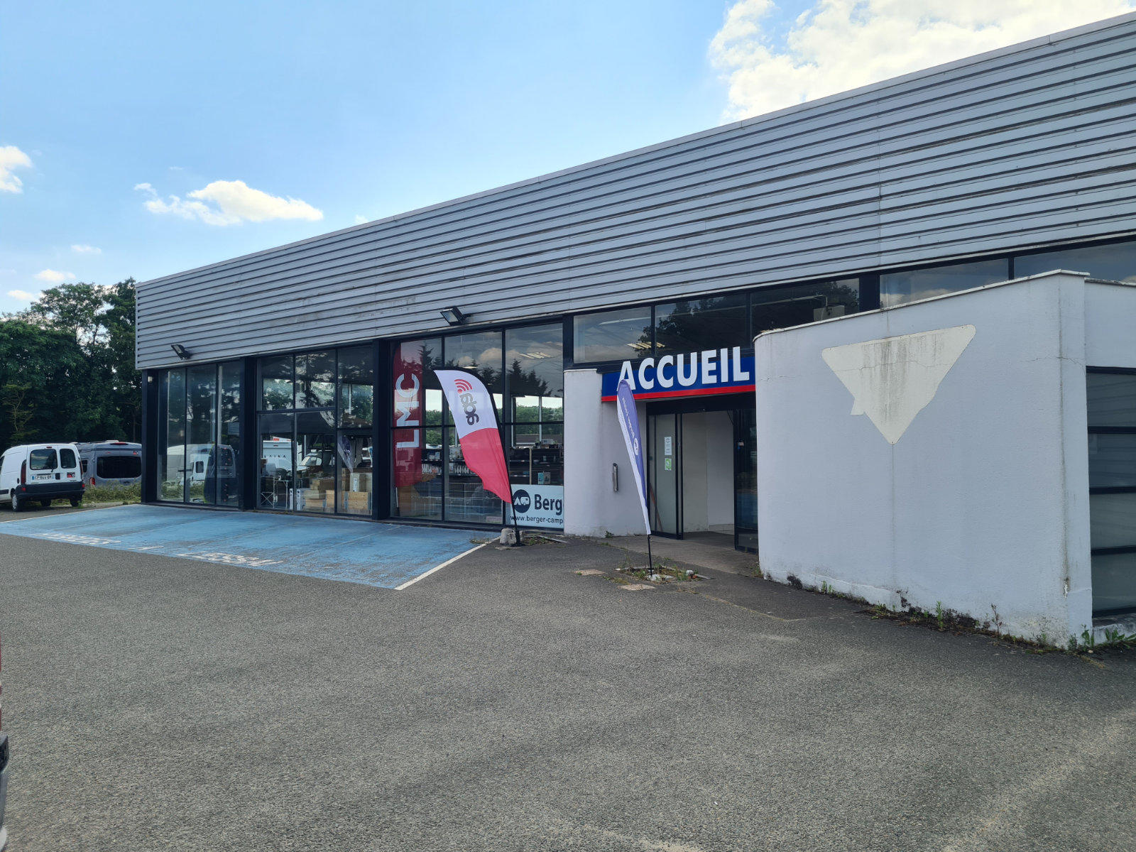 Au&szlig;enansicht eines niedrigen Gesch&auml;ftsgeb&auml;udes mit Glasfront, Schild 'ACCUEIL' &uuml;ber dem Eingang, zwei Werbefahnen neben der T&uuml;r, blau markierter Parkplatz, wei&szlig;es Fahrzeug links, gepflasterter Parkplatz, bew&ouml;lkter Himmel