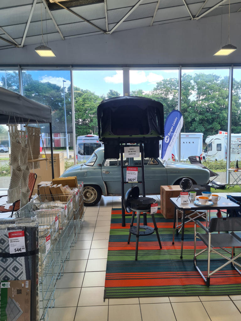 Hellblaues Auto mit ge&ouml;ffneter Motorhaube vor gro&szlig;en Fenstern; im Vordergrund Tische, St&uuml;hle, Kartons und gestreifte Teppiche