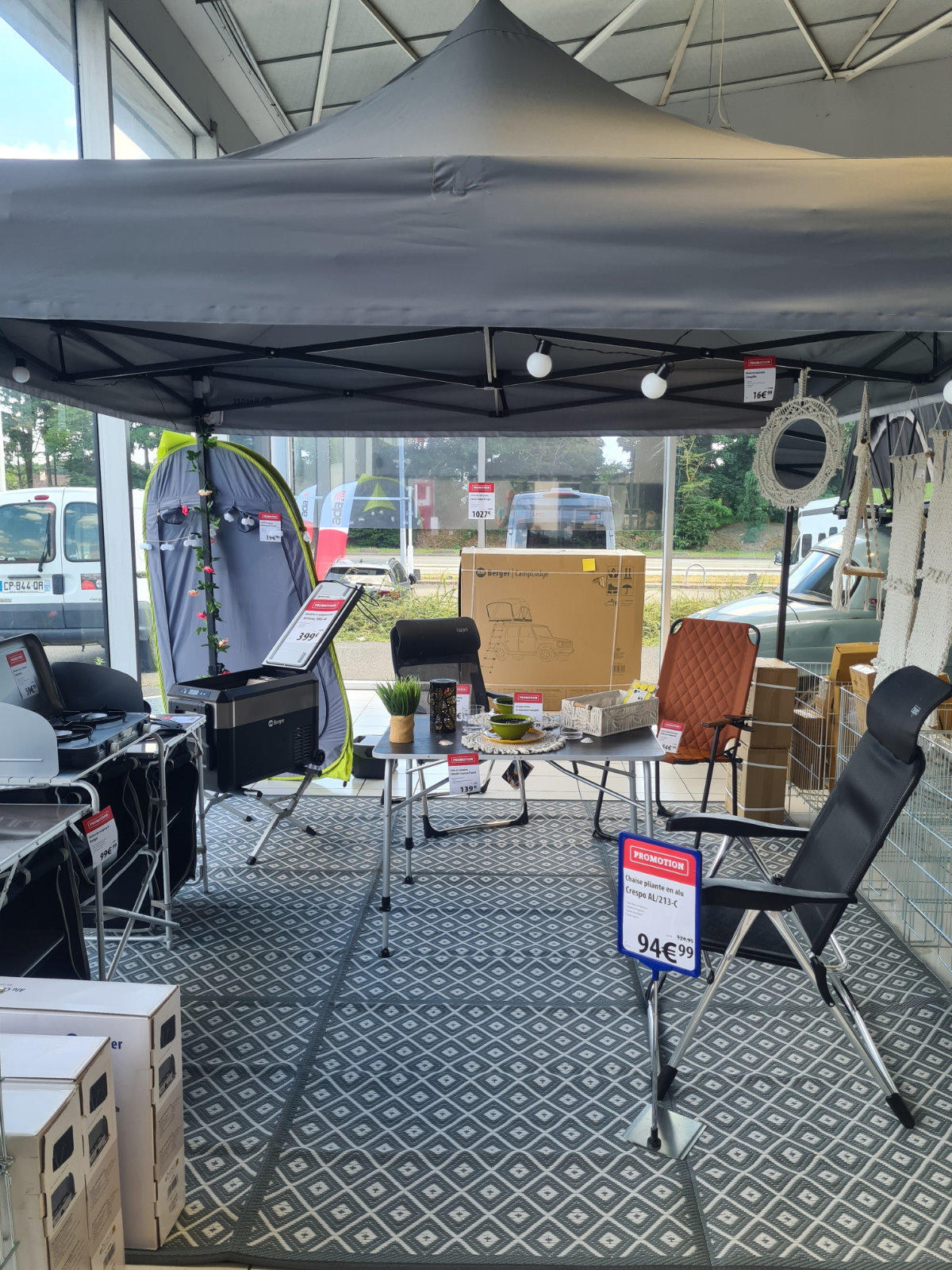 Schwarzes Faltzelt mit spitzem Dach &uuml;ber Teppich mit Diamantmuster, darunter Tisch mit Karton und Pflanze, St&uuml;hle, h&auml;ngende Lampen und Preisschilder; durch Fenster im Hintergrund parkende Fahrzeuge sichtbar
