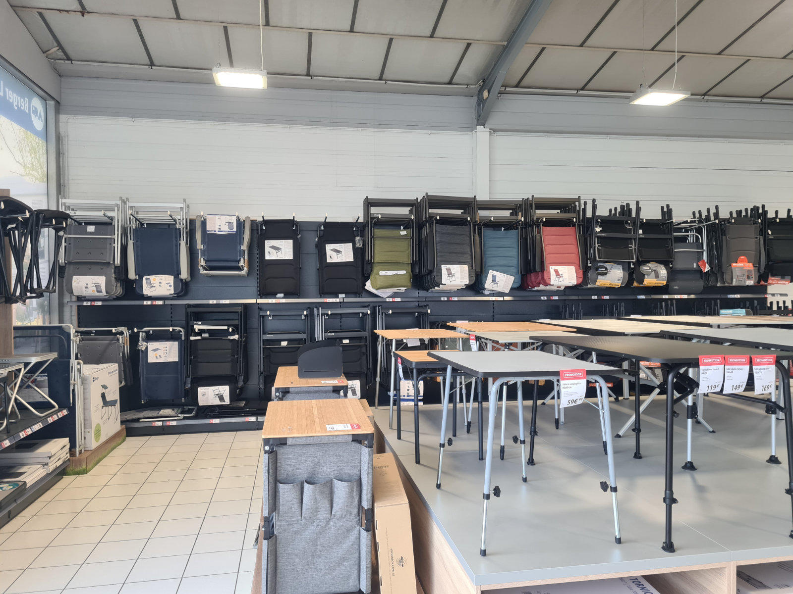 Verkaufsraum mit Regalen voller gestapelter St&uuml;hle und zusammengeklappter Tische, Ausstellungs&shy;tische auf Podesten vorn, gefliester Boden und Deckenleuchten