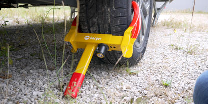 Fahrzeugrad mit gelb-roter Radkralle auf kiesigem Boden, rechts Teil eines blauen Jeansbeins sichtbar