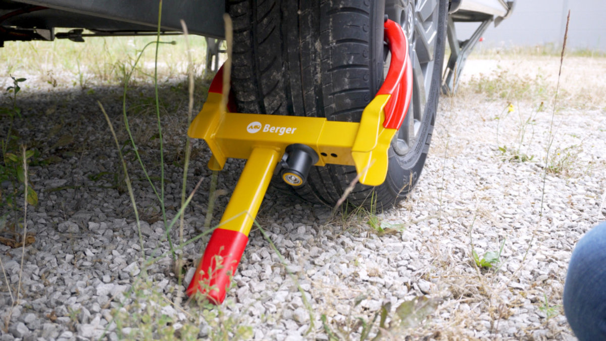 Fahrzeugrad mit gelb-roter Radkralle auf kiesigem Boden, rechts Teil eines blauen Jeansbeins sichtbar