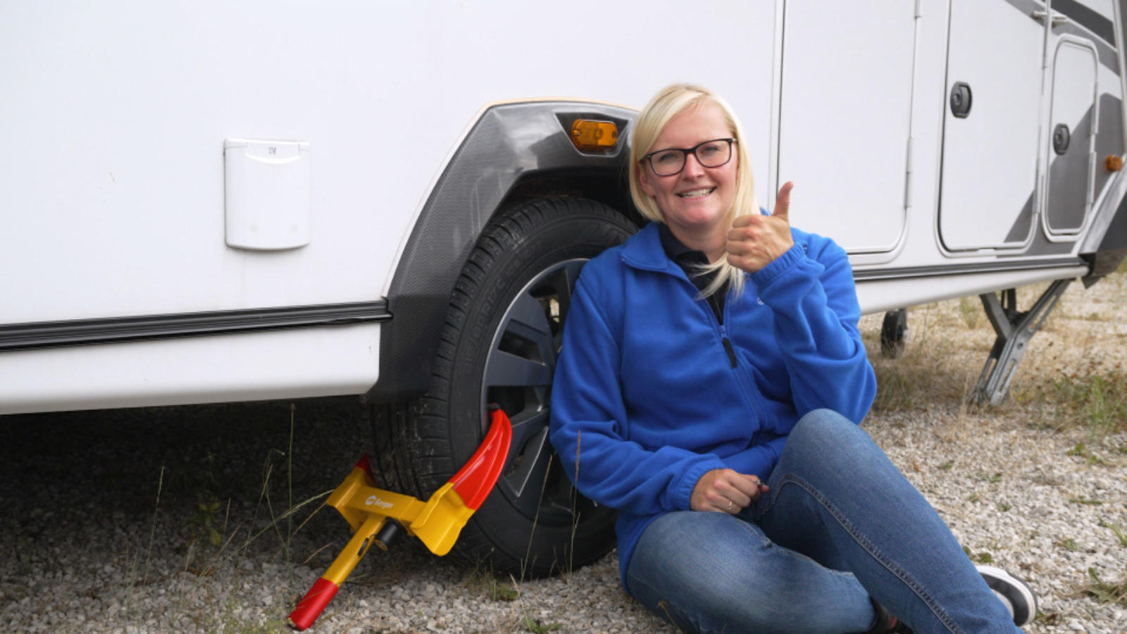 Person sitzt auf Kies neben einem weißen Wohnwagenrad, rote-gelbe Radkralle am Reifen, trägt blaue Jacke, Jeans und Brille, zeigt Daumen hoch