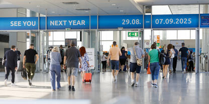 Menschen gehen durch eine Flughafenhalle, einige ziehen Gep&auml;ck; &uuml;ber ihnen blaue Anzeigetafeln mit 'SEE YOU', 'NEXT YEAR' und den Daten '29.08.' und '07.09.2025'