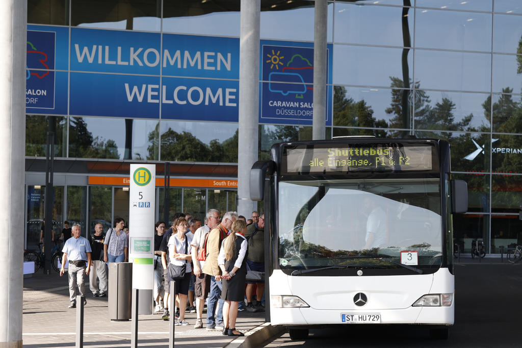 Bus an einer Haltestelle vor einem Glasgeb&auml;ude mit gro&szlig;em 'WILLKOMMEN WELCOME'-Schild; mehrere wartende Personen an der Haltestelle