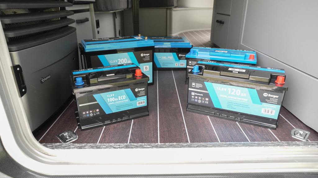 Mehrere schwarze rechteckige Batterien mit blauen Etiketten auf dunklem Holzfußboden im geöffneten Laderaum eines Fahrzeugs