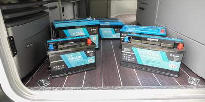 Mehrere schwarze rechteckige Batterien mit blauen Etiketten auf dunklem Holzfu&szlig;boden im ge&ouml;ffneten Laderaum eines Fahrzeugs