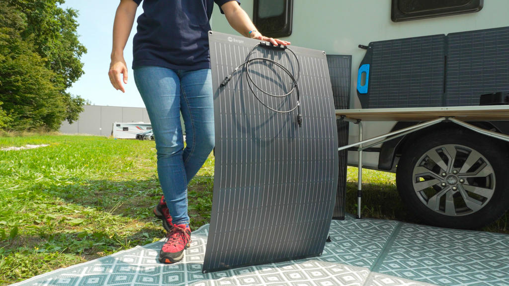 Unterkörper einer Person in Jeans und roten Schuhen, die neben einem Wohnwagenrad eine große gerillte dunkelgraue Matte mit aufgewickeltem Kabel hält; gemusterter Bodenbelag und Gras im Hintergrund