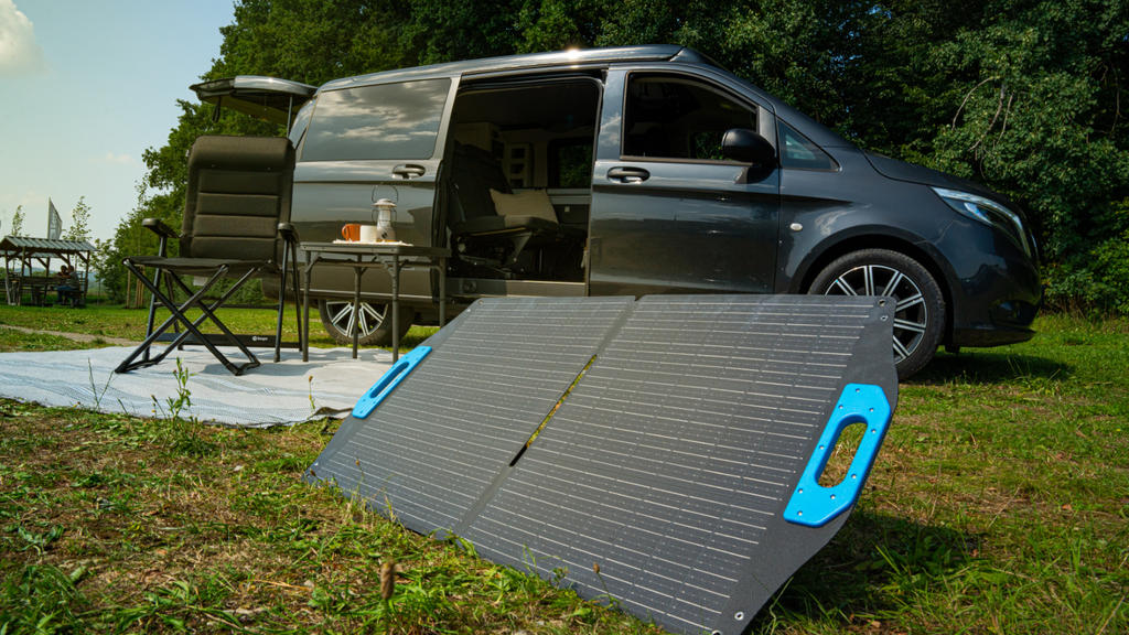Faltbares Solarpanel mit blauen Griffen auf Gras vor einem geparkten grauen Camper-Van mit offener Schiebetür, Klappstuhl, kleinem Tisch und heller Bodenmatte; Bäume im Hintergrund
