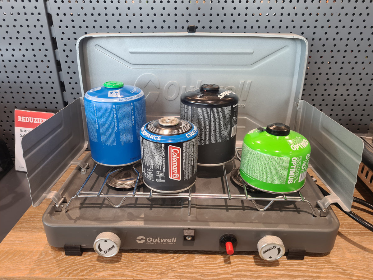 Portabler Camping-Gaskocher mit Windschutz, drei Gaskartuschen (blau, schwarz, gr&uuml;n) auf dem Kochrost, Holzfl&auml;che, Lochwand im Hintergrund