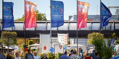 Menschen auf Fu&szlig;weg unter f&uuml;nf bunten Fahnen vor Geb&auml;ude mit &uuml;berdachtem Durchgang; Verkehrsschilder und B&auml;ume