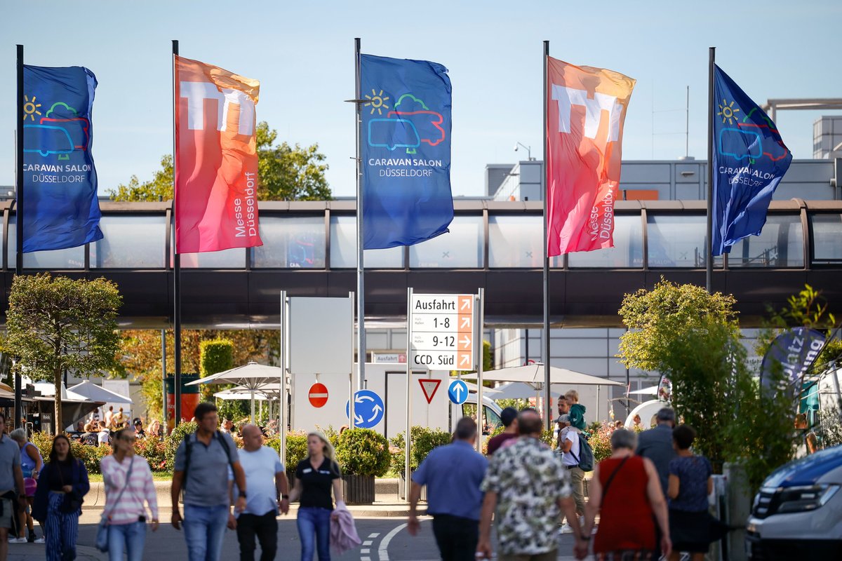 Menschen auf Fu&szlig;weg unter f&uuml;nf bunten Fahnen vor Geb&auml;ude mit &uuml;berdachtem Durchgang; Verkehrsschilder und B&auml;ume
