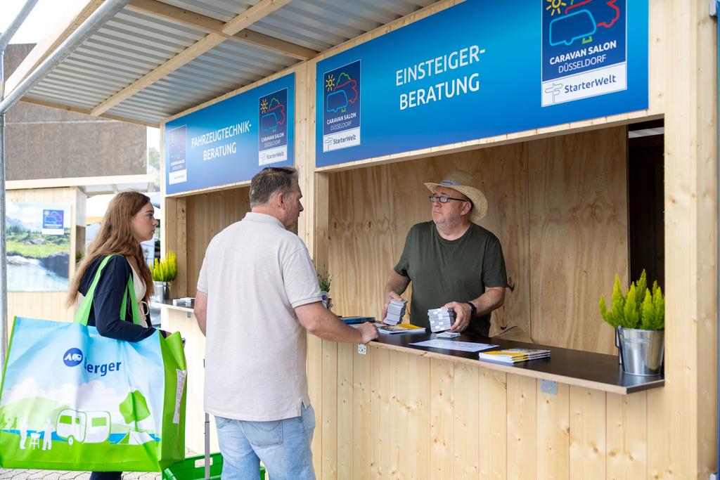 Drei Personen an einem hölzernen Außentresen; eine Person hinter dem Tresen mit Hut und grünem T‑Shirt, zwei Personen davor, eine in beigem Hemd, eine mit großer grün‑blauer Tragetasche; blauer Schild über dem Tresen mit der Aufschrift EINSTEIGER-BER