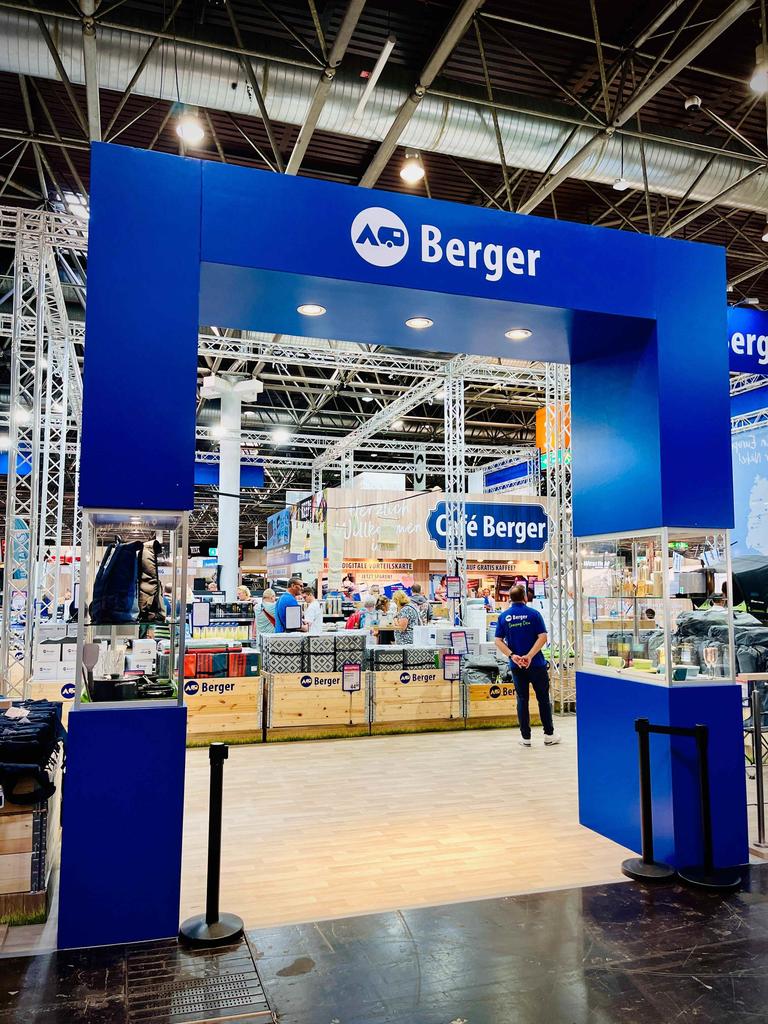 Blauer rechteckiger Eingang mit dem Schriftzug 'Berger' und rundem Logo; dahinter Glasschauvitrinen, Verkaufstheken, weitere Stände und Personen in einer Messehalle