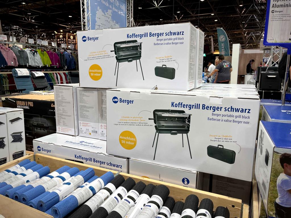 Gestapelte Kartons mit Aufschrift "Koffergrill Berger schwarz" und Abbildung eines Grills; im Vordergrund aufgerollte Textilien in Blau, Weiß und Schwarz; Regale und weitere Produkte im Hintergrund; Personen sichtbar