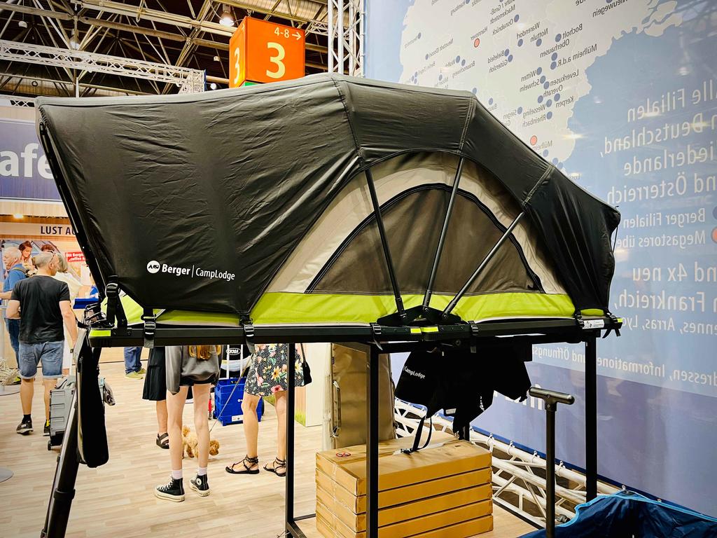 Schwarzes und grünes aufgebautes Dachzelt auf schwarzem Metallgestell in einer Messehalle; Holzkiste darunter, orange Zahl 3 im Hintergrund, mehrere Personen im Vorbeigehen