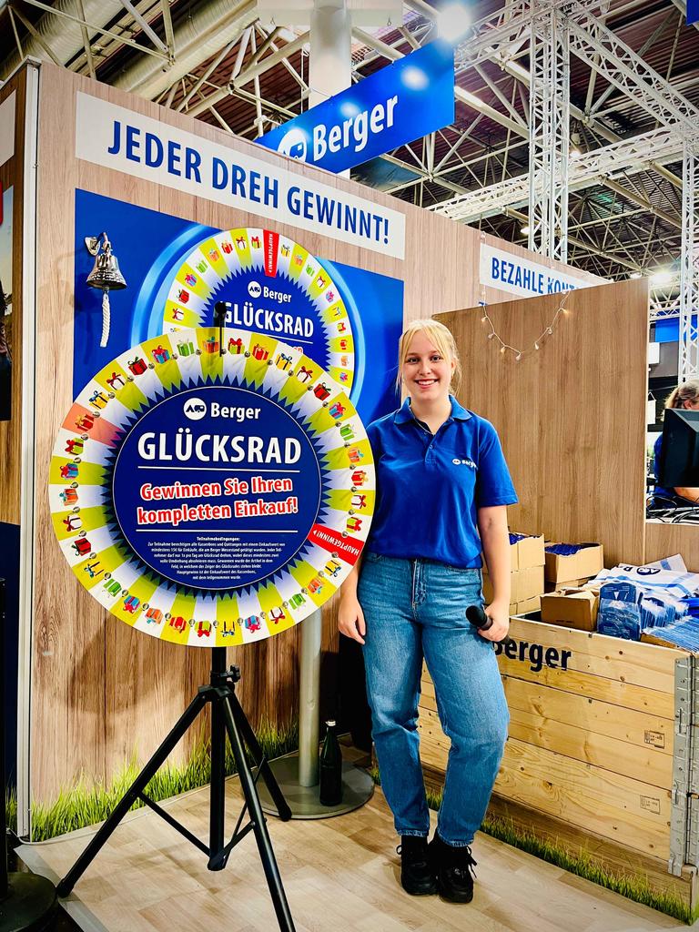 Person in blauem Poloshirt und Jeans steht neben einem bunten Glücksrad auf Stativ; im Hintergrund Holzverkleidung, Kisten, ein Schild mit der Aufschrift "JEDER DREH GEWINNT!" und ein "Berger"-Logo