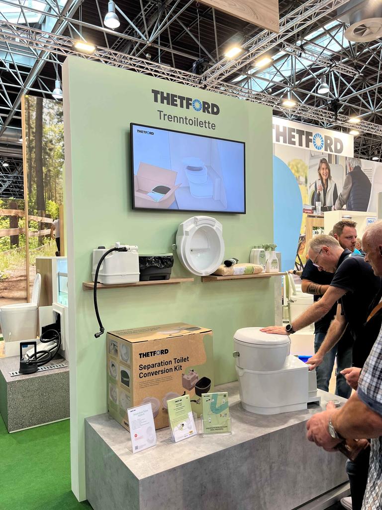 Messestand mit grüner Rückwand und Schrift "THETFORD", Bildschirm zeigt Toiletten, offene Toilettenbrille auf Theke, Produktkarton auf Podest, mehrere Personen rechts