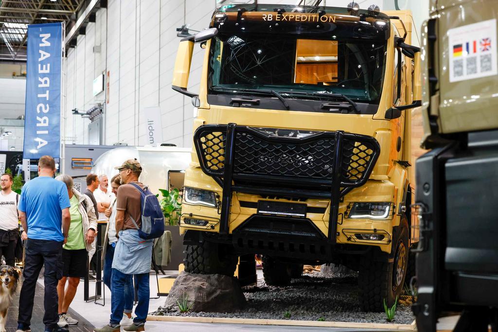 Großer gelber Lkw mit verstärktem Frontschutz in einer Messehalle, Personen stehen links daneben