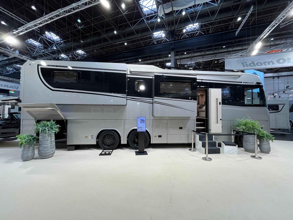 Großes graues Wohnmobil in einer Ausstellungshalle, geöffnete Seitentür mit Trittstufen, Pflanzenkübel und Informationsständer neben dem Fahrzeug, sichtbare Hallendecke mit Beleuchtung, keine Personen