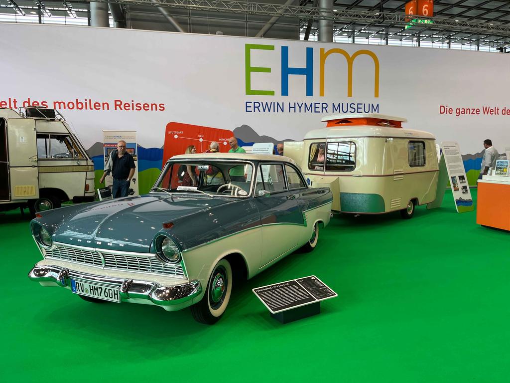 Zweifarbiger grüner und weißer Pkw auf grünem Boden vor einem cremegrünen Wohnwagen, Messewand mit der Aufschrift 'EHM Erwin Hymer Museum', weitere Wohnmobile und mehrere Personen im Hintergrund