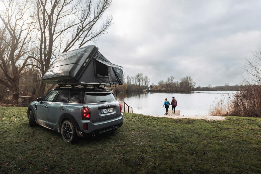 Auto mit aufgestelltem Dachzelt auf einer Wiese neben einem See; zwei Personen gehen am Ufer; B&auml;ume und bew&ouml;lkter Himmel